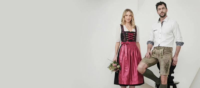Woman in black dirndl with pink apron and man in lederhosen, traditional Oktoberfest outfits.