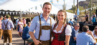 Couple wearing traditional dirndl and lederhosen enjoying an Oktoberfest event in Arizona with desert scenery and live music.
